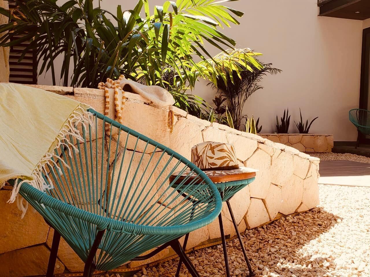 A hammock chair and a small table with a cushion in a sunny outdoor area with plants and a stone wall.