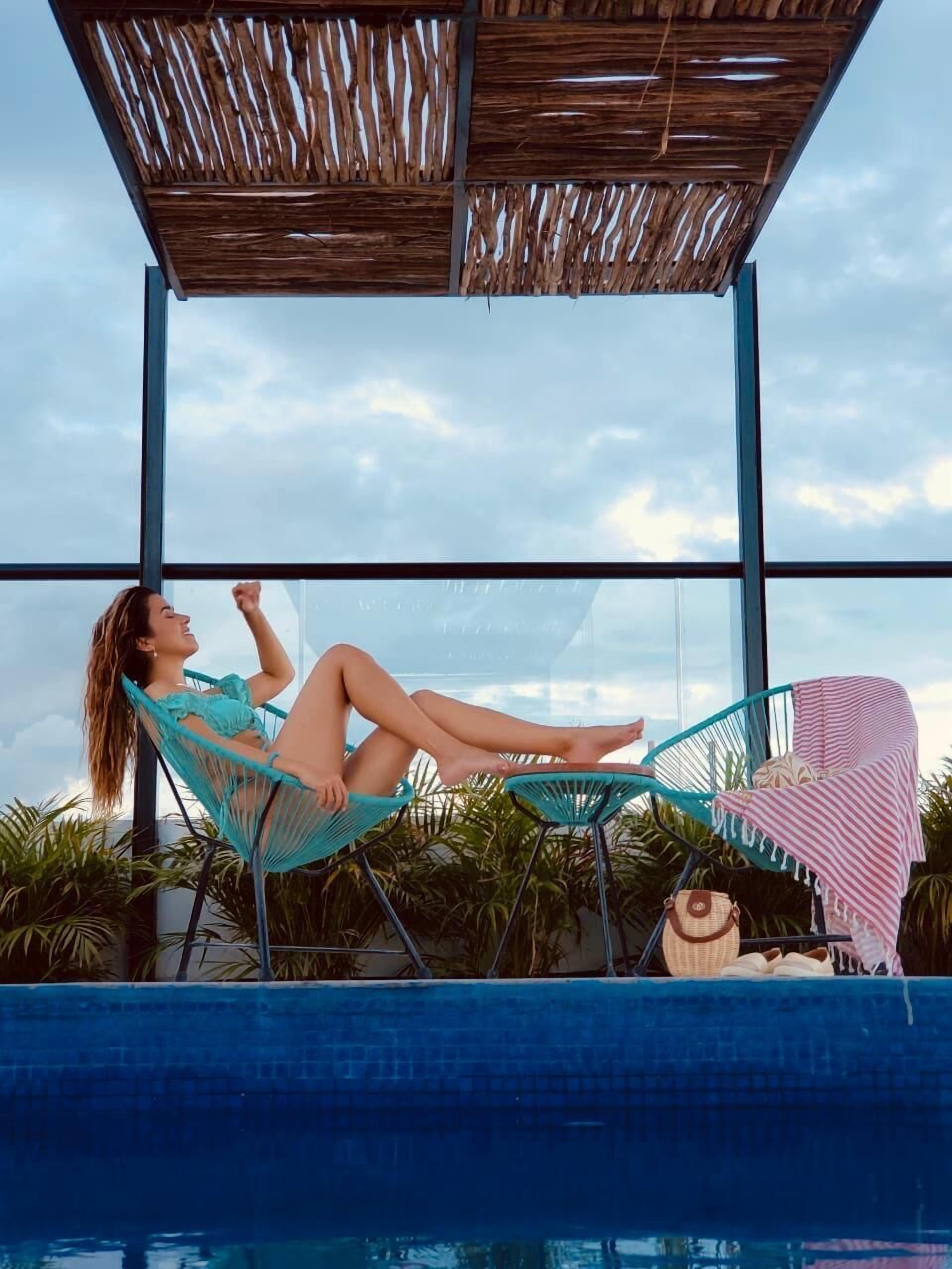 Une personne se détend dans un fauteuil suspendu près d’une piscine, avec une vue sur le ciel à travers de grandes fenêtres.