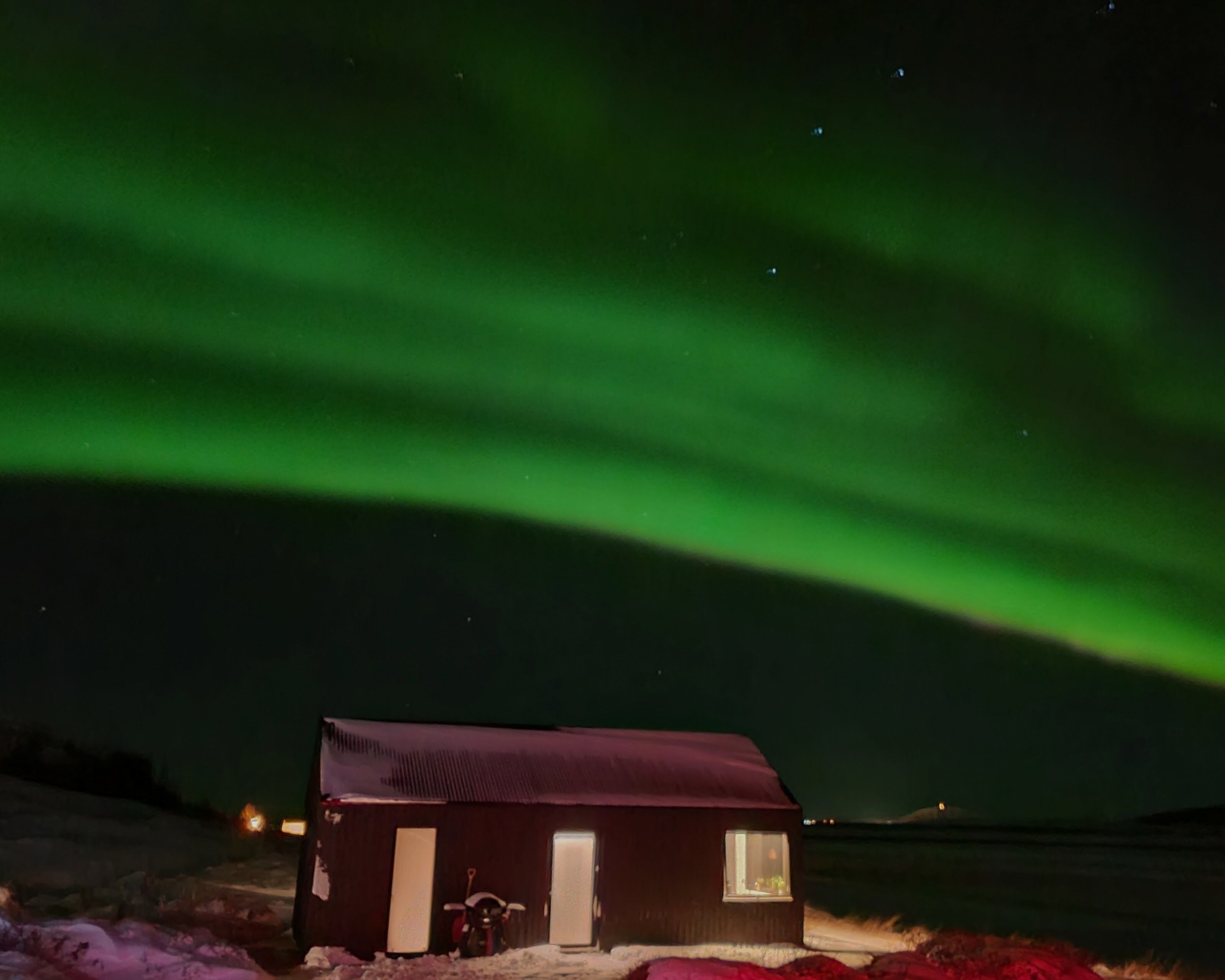 Une petite cabane de couleur sombre, dotée d’une seule fenêtre visible, située dans un paysage enneigé, sous un ciel nocturne parsemé d’étoiles.