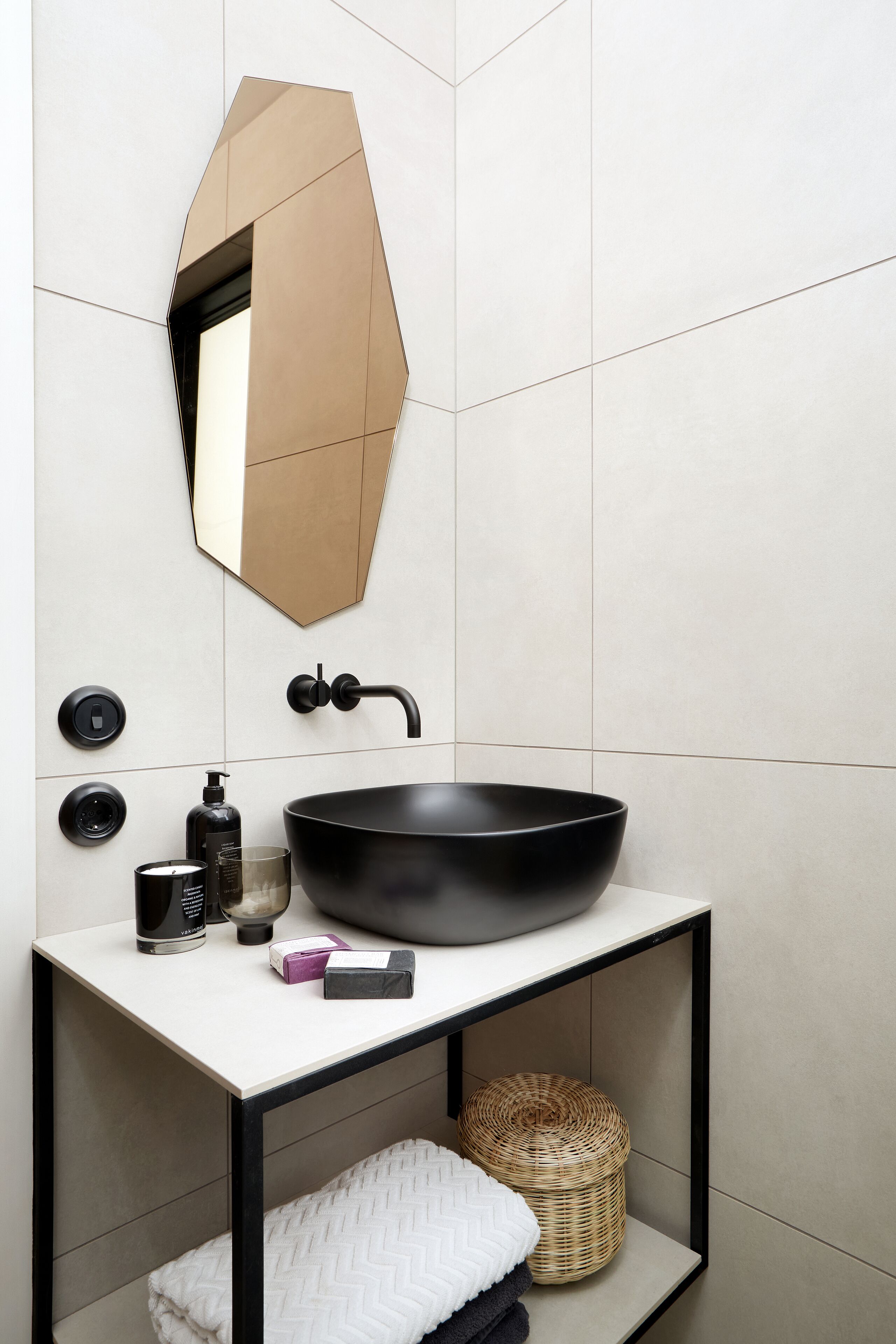 A modern bathroom with a large black sink, a geometric mirror, and a small shelf with toiletries.