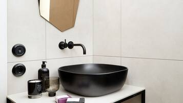 A modern bathroom with a large black sink, a geometric mirror, and a small shelf with toiletries.
