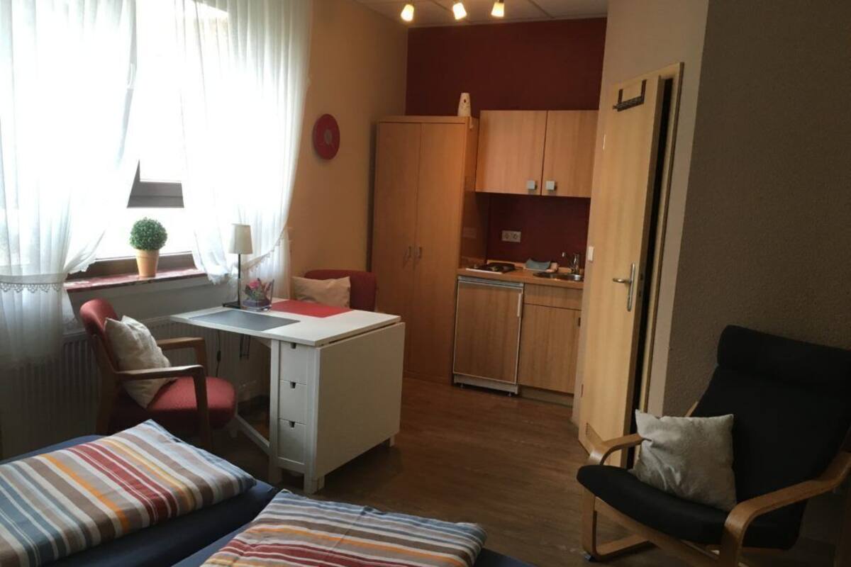 Une pièce de taille modeste comprenant un lit, un bureau, une chaise, un petit coin cuisine et une fenêtre avec des rideaux.