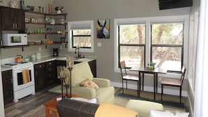 Una cocina moderna con microondas, estufa y diversos utensilios de cocina en estantes. Hay una zona de comedor con mesa y sillas, y un sofá.