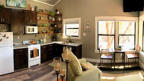 Cocina con armarios de madera oscura, un refrigerador blanco y un microondas. Hay un comedor con mesa y sillas, y una ventana con vista a árboles.