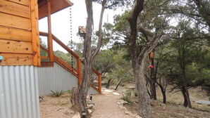 Una cabaña de madera con revestimiento metálico, un camino de grava que conduce a una terraza de madera, y árboles circundantes.