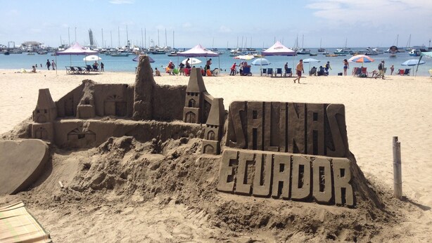 Una spiaggia con un castello di sabbia con la scritta "COSTA ECUDOR" e un porto turistico sullo sfondo.