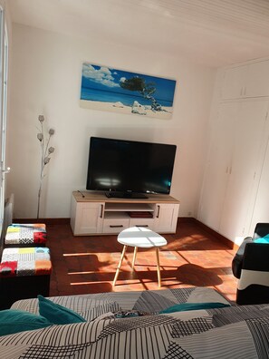 Un salon avec un téléviseur à écran plat posé sur un meuble en bois, une petite table blanche, des coussins à motifs et un tableau représentant un arbre accroché au mur.