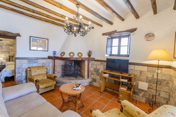A cozy living room with a stone fireplace, wooden beams, a television on a wooden stand, and a comfortable seating area. - Casa Rural El Pilarillo (Alcaucín)