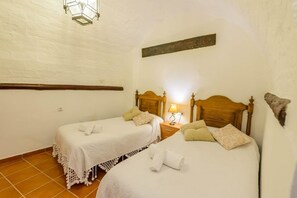 A room with two beds, each with a wooden headboard, a nightstand with a lamp, and a white wall with a wooden beam. - Casa Rural El Pilarillo (Alcaucín)