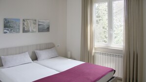 Una cama doble con edredón blanco y morado, dos almohadas blancas y una mesita de noche con lámpara. Encima de la cama, hay tres cuadros enmarcados en la pared.