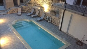 A rectangular swimming pool with a stone border, surrounded by lounge chairs and a stone wall.
