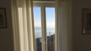 A room with a sliding glass door leading to a balcony with a stone floor and a view of the sea.