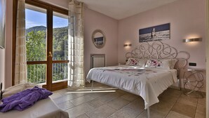 Un dormitorio con cama, mesita de noche, silla, ventana con cortinas, radiador y un cuadro en la pared.