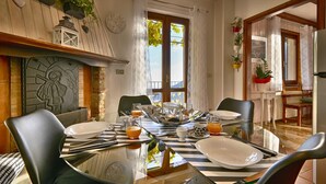 Un comedor con una mesa de vidrio dispuesta para cuatro personas, rodeada de sillas. Se observa una pared de madera con un reloj y detalles decorativos, además de un ventanal con cortinas.