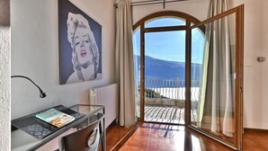 A room with a glass table, a chair, a radiator, a painting of a woman, and a view of a beach and mountain through a glass door. - Armonia by Theater Home, with two parking spaces and beautiful lake view (Tignale)