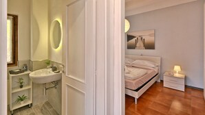 A modern bedroom with a bed, bedside table, and a nightstand. There is a bathroom with a sink, mirror, and shelves. - Armonia by Theater Home, with two parking spaces and beautiful lake view (Tignale)