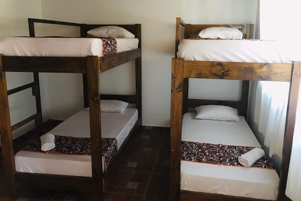 Duas camas beliches com roupa de cama branca e mantas estampadas, estrutura de madeira e chão ladrilhado.