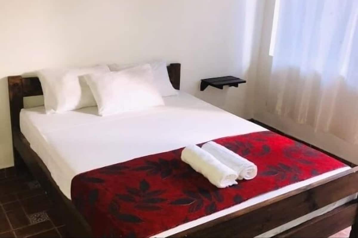 Uma cama bem arrumada com lençóis brancos e uma colcha floral vermelha, uma mesinha de cabeceira e uma janela com cortinas leves.
