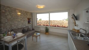 A modern kitchen with a stone wall, a dining area with a table set for two, and a large window with a view of a tiled roof. - Menèk by Theater Home, in the historic centre (Tignale)