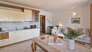 A modern kitchen with white cabinets, a dining area with a table set for two, and a living room with a sofa and a painting on the wall. - Michela 4  by Theater Home, with private garden and lake view (Tignale)