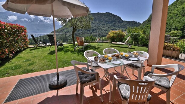 An outdoor patio with a table set for breakfast, surrounded by chairs, and an umbrella providing shade. - Michela 4  by Theater Home, with private garden and lake view (Tignale)