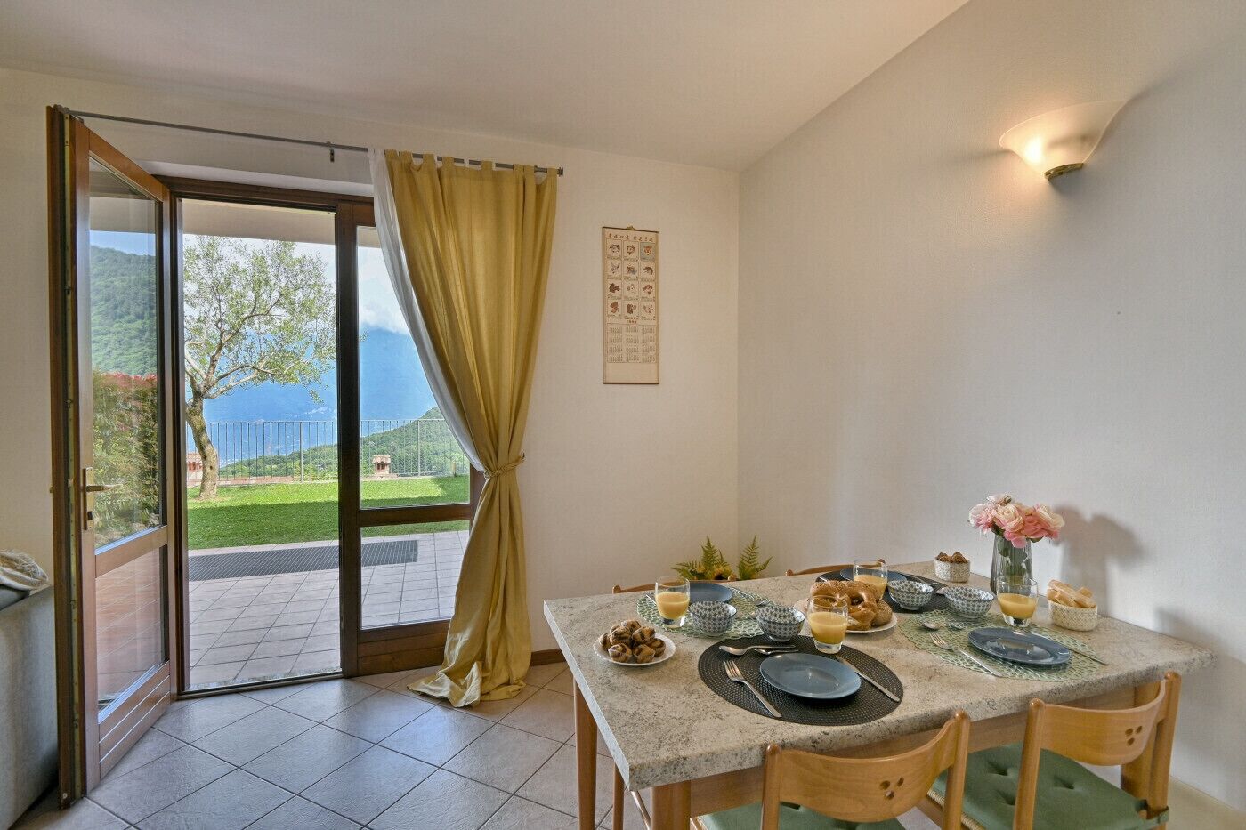 A dining area with a table set for two, a view of a mountainous landscape through a glass door, and a wall-mounted light fixture.