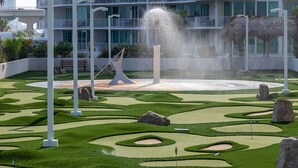 Un jardín arbolado con una fuente, columnas blancas y un edificio con balcones al fondo.