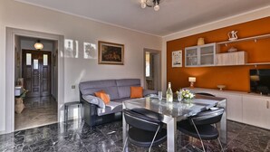 Un salon moderne avec un canapé gris, des chaises noires, une table à manger en verre et un coin cuisine doté de placards et d’un téléviseur.