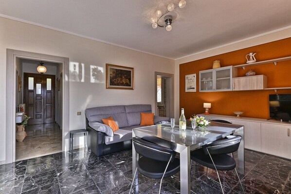 A modern living room with a grey sofa, black chairs, a glass dining table, and a kitchen area with cabinets and a TV.