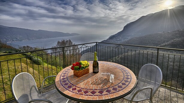 Une table ronde avec une bouteille et des fleurs, placée sur un balcon donnant sur un lac et des montagnes.