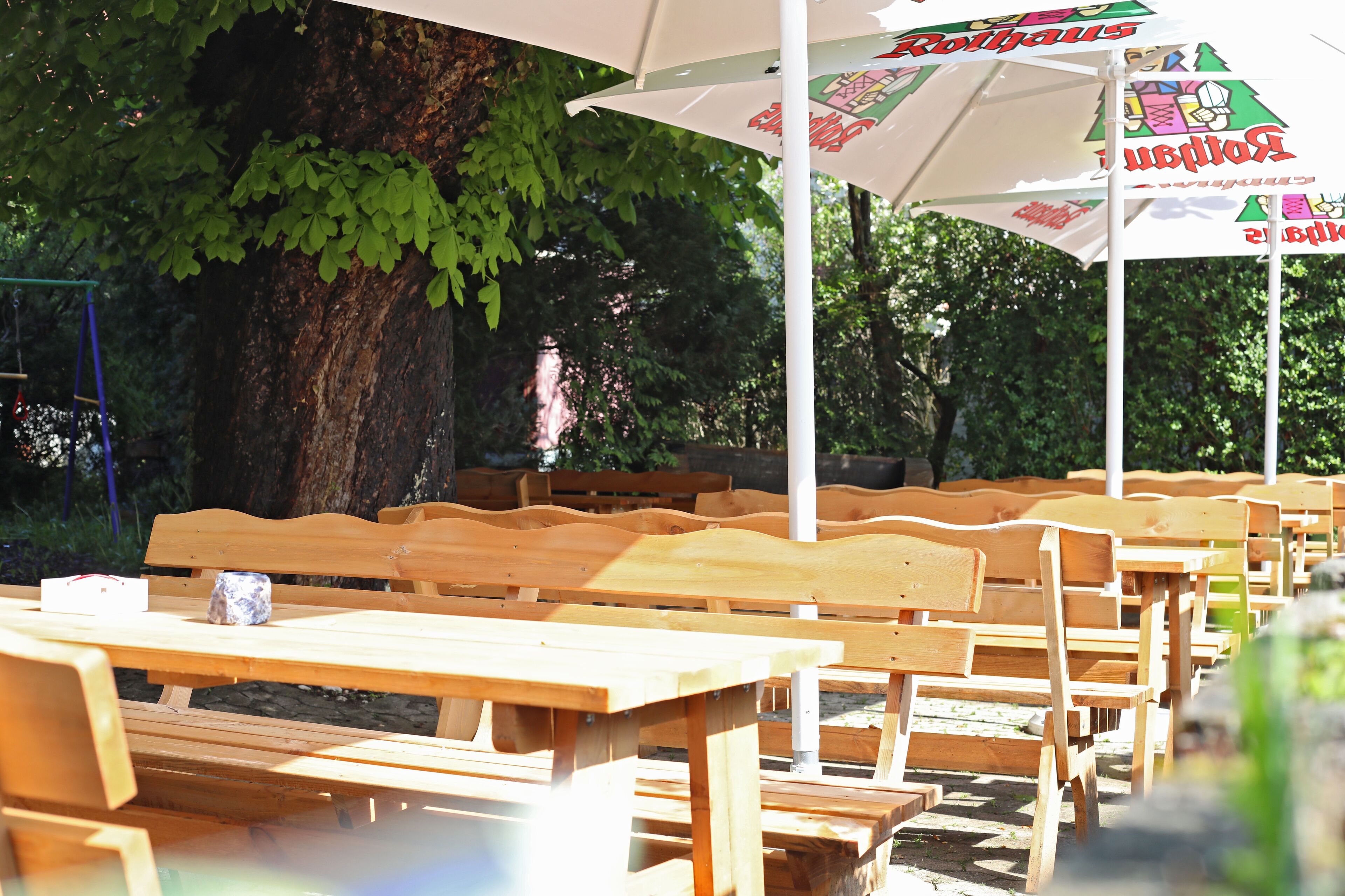 Des bancs et des tables de pique-nique en bois sont disposés en rangées à l’extérieur, avec un grand arbre et des parasols en arrière-plan.