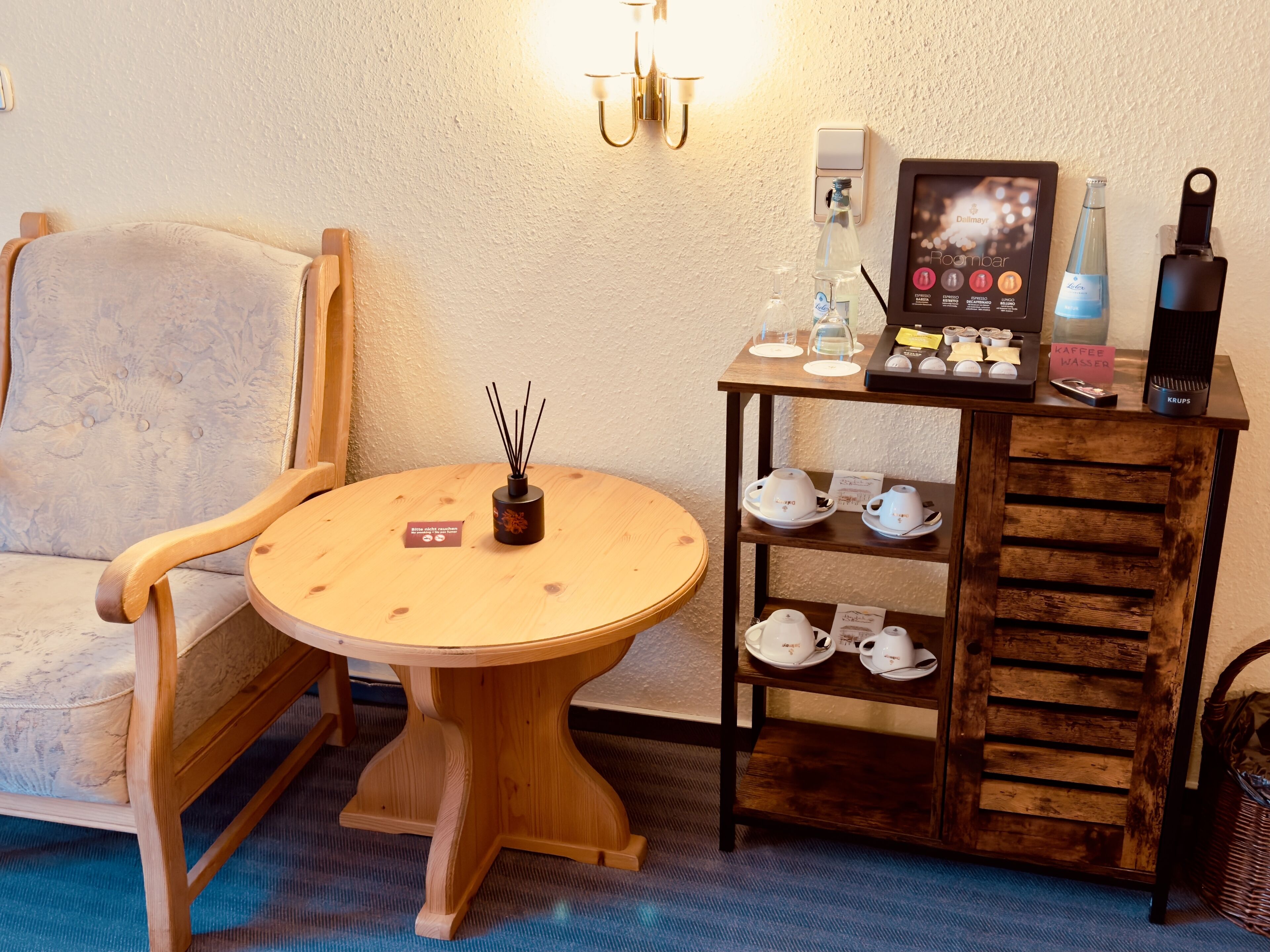 Eine gemütliche Sitzecke mit einem Holzstuhl und einem kleinen runden Tisch, einem Regal mit Teetassen und einer Kaffeemaschine, sowie einer wandmontierten Lampe.