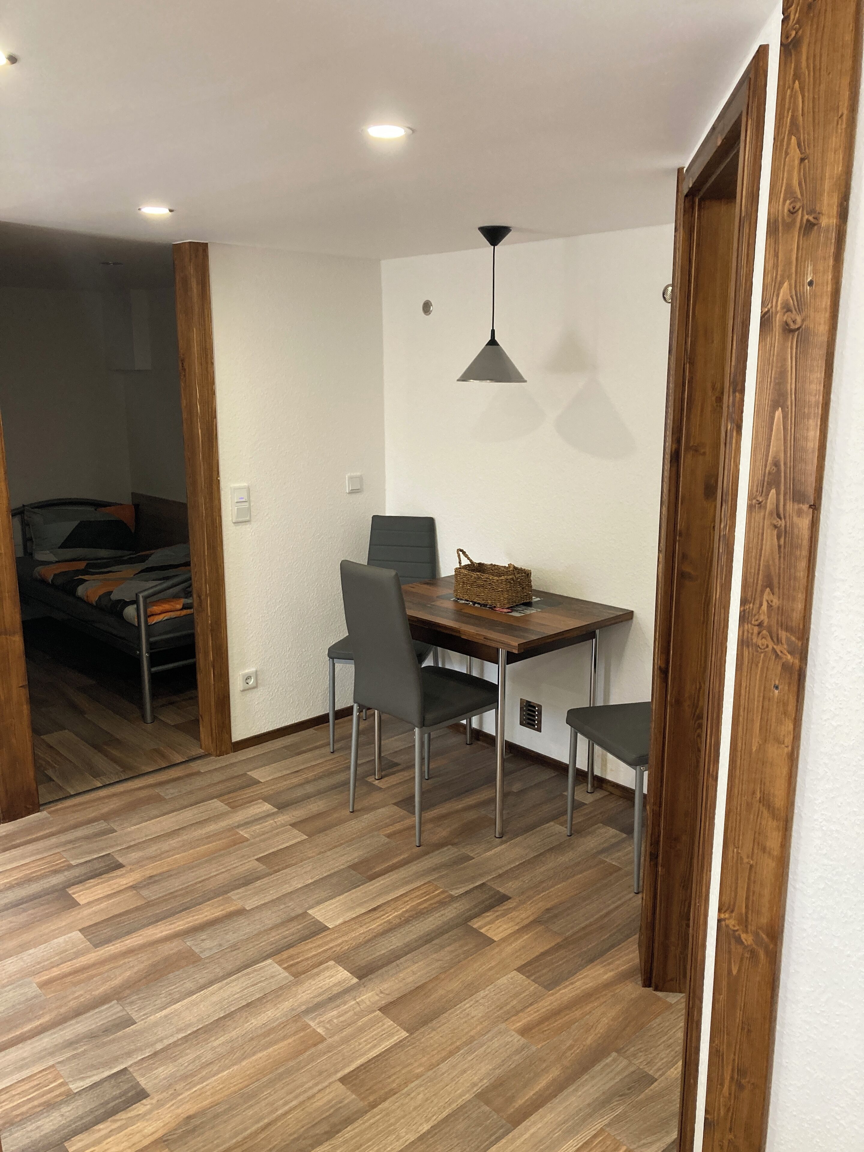 Ein kleines Zimmer mit Holzboden, einem Esstisch mit Stühlen, einer Pendelleuchte und einem Bett im Hintergrund.