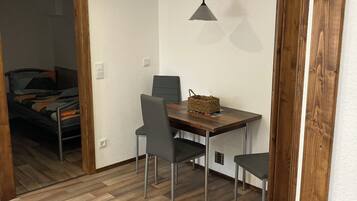 Ein kleines Zimmer mit Holzboden, einem Esstisch mit Stühlen, einer Pendelleuchte und einem Bett im Hintergrund.