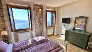 Une chambre à coucher avec un lit, une commode, une chaise et un miroir. Il y a deux fenêtres avec des rideaux, et on peut voir un mur en pierre.
