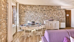 Une pièce aux murs de pierre, avec une table à manger dressée pour quatre personnes, un buffet blanc et un canapé violet.