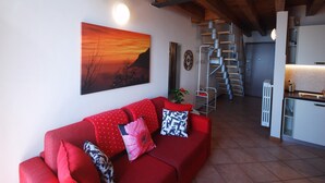 A living room with a red sofa, a painting of a sunset, a spiral staircase, and a kitchen area. - Lince by Theater Home, with pool and lake view (Tignale)