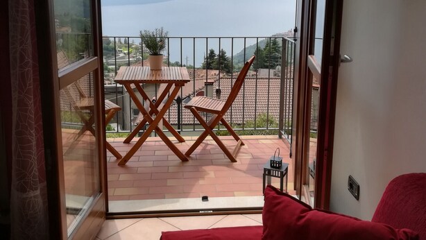 A room with a sliding glass door leading to a balcony with a table and chairs, a red sofa, and a view of the sea and mountains. - Lince by Theater Home, with pool and lake view (Tignale)