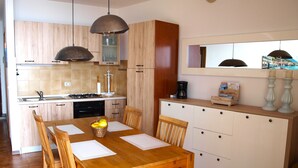 Een compacte keuken met houten kastjes, een eettafel met stoelen, een koffiezetapparaat en een klok aan de muur.