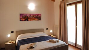 Una habitación de hotel con una cama, una mesita de noche, una lámpara, un cuadro de un paisaje urbano y una ventana con cortinas.