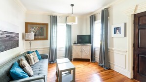 A living room with a sofa, a coffee table, a TV on a cabinet, and a door. - Spacious Cottage w/ King Bed, Patio, Easy Parking by Lucky Savannah (Savannah)