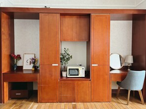 A compact room with a wooden desk, a chair, a microwave, and a small cabinet with a mirror and lamp.