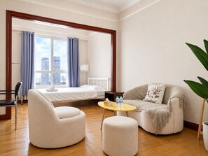 A hotel room with a bed, two armchairs, a small table, a view of the city, and a plant.