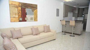 Un salon moderne avec un canapé beige, des coussins à motifs, un grand tableau abstrait et un coin cuisine doté d’un tabouret de bar.