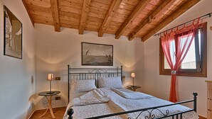 Une chambre avec un plafond en bois, un lit recouvert d’une literie à motifs floraux, deux tables de chevet avec des lampes et une fenêtre avec un rideau.