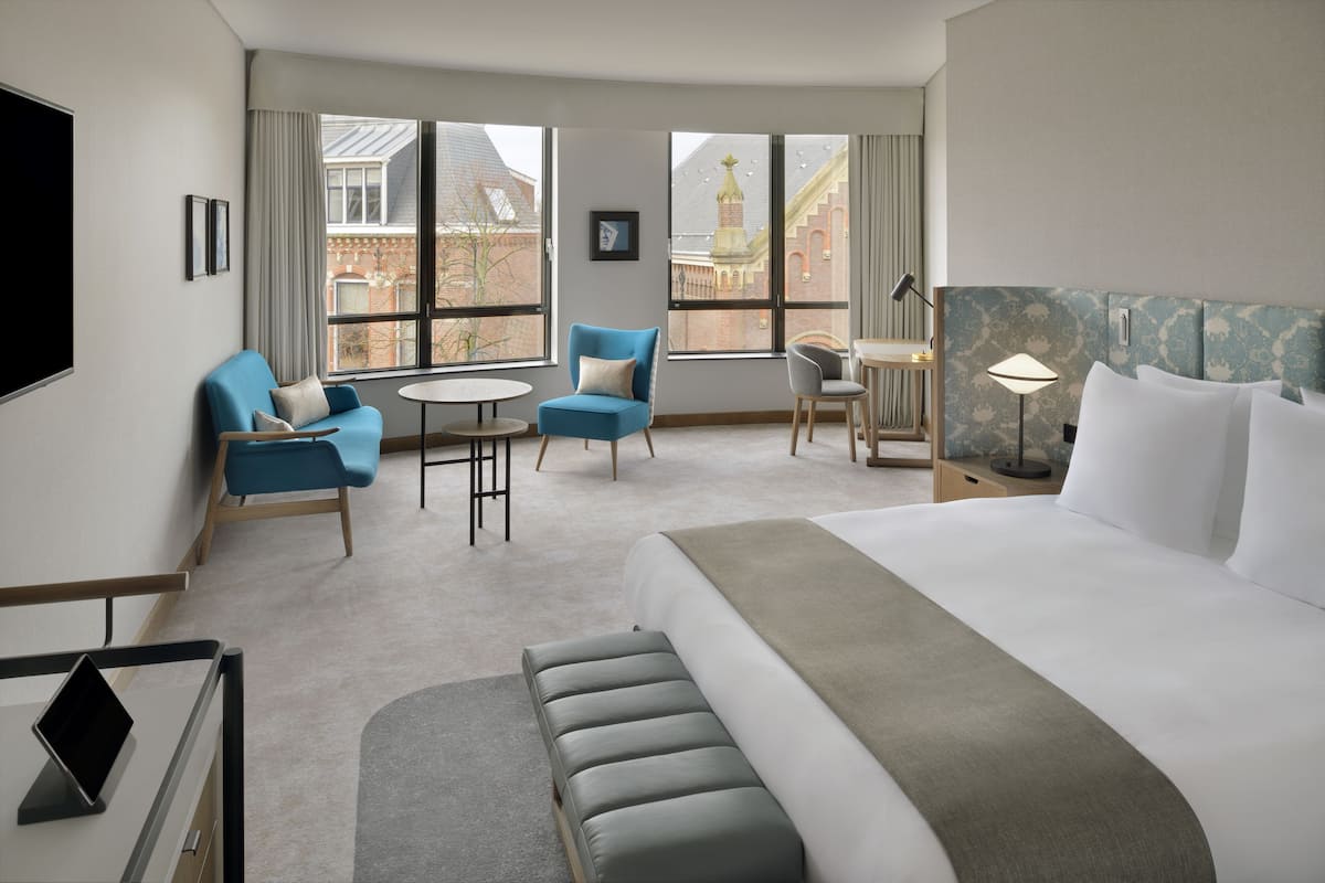 A hotel room with a large bed, a seating area with chairs and a table, a desk with a laptop, and large windows offering a view of a building with a tower.