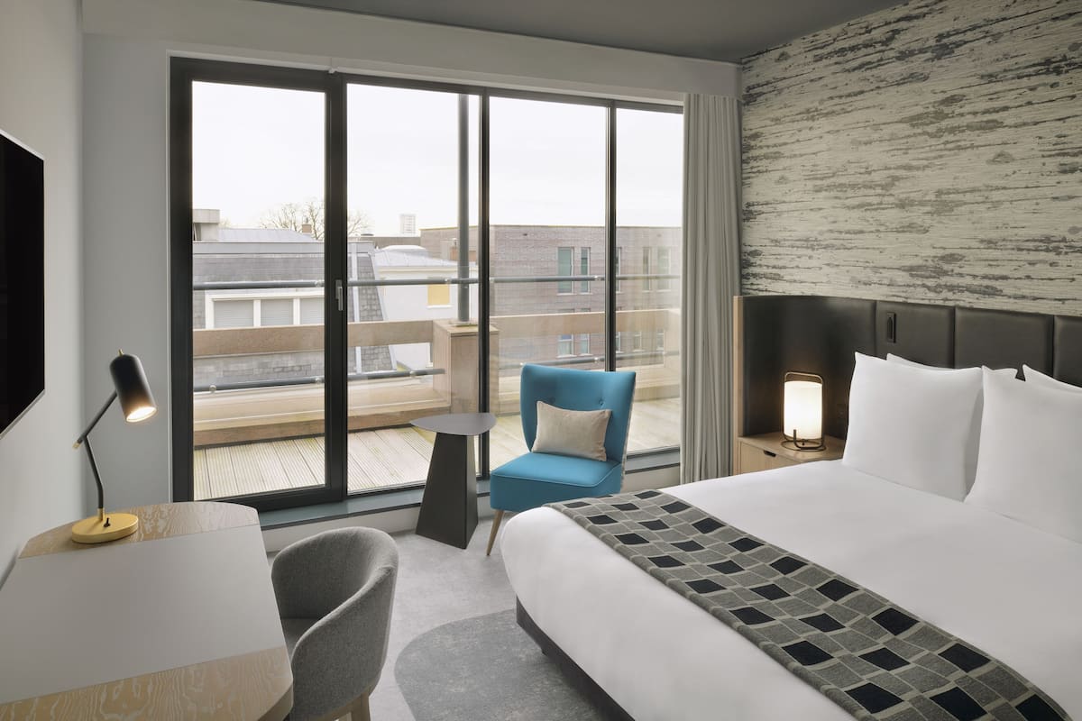 A hotel room with a large bed, a desk, a chair, a small table, and a balcony with a view of buildings.