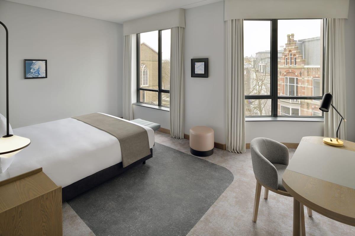 A hotel room with a bed, a desk, a chair, and two windows.