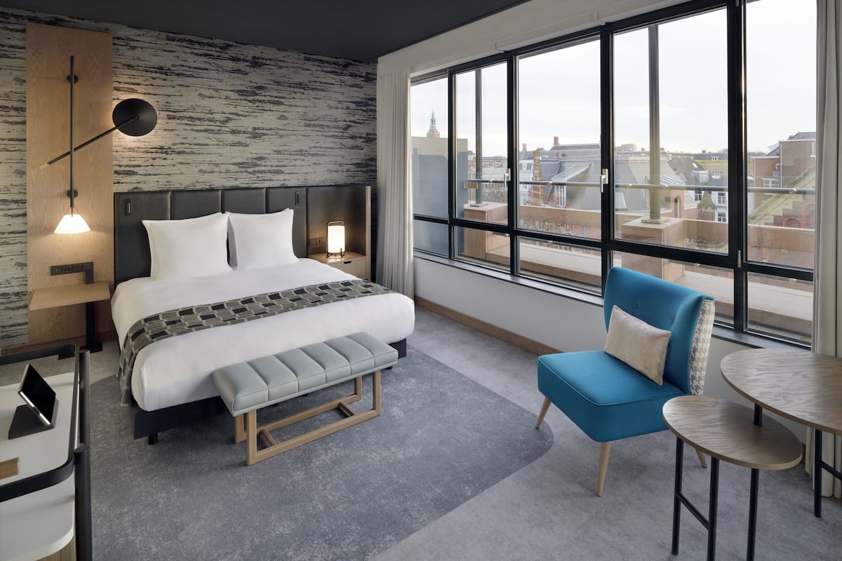 A modern hotel room with a large bed, a blue armchair, a small table, and a view of the city through large windows.