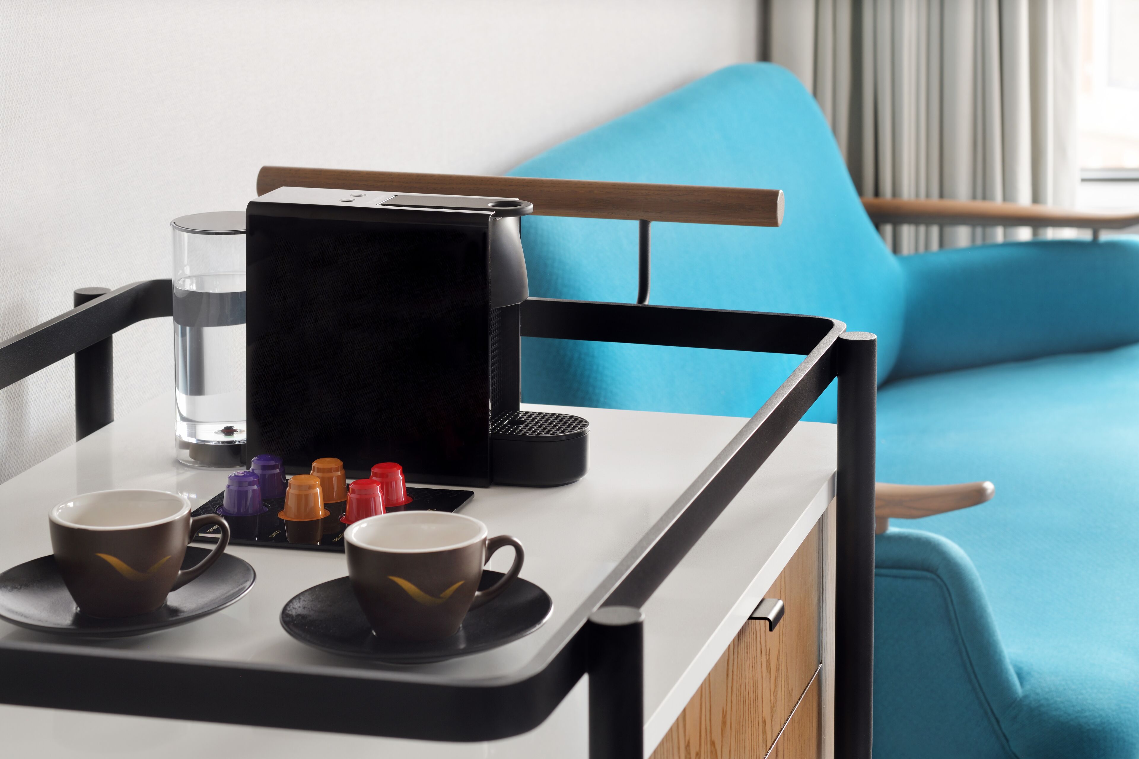 A compact kitchenette with a coffee maker, two cups, and a glass of water on a tray.